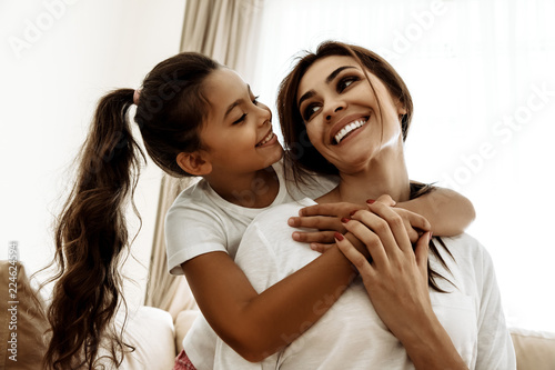 Family. Love. Mom and daughter are hugging, looking at each other and smiling while sitting on couch at home