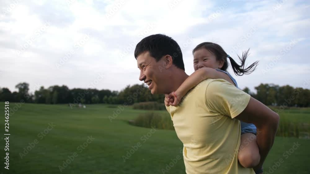 Cheerful asian father giving his cute laughing preschool daughter piggyback ride while having fun in summer park. Adorable little chinese girl enjoying piggyback ride with her smiling dad outdoors. Cheerful asian father giving his cute laughing preschool daughter piggyback ride while having fun in summer park. Adorable little chinese girl enjoying piggyback ride with her smiling dad outdoors.