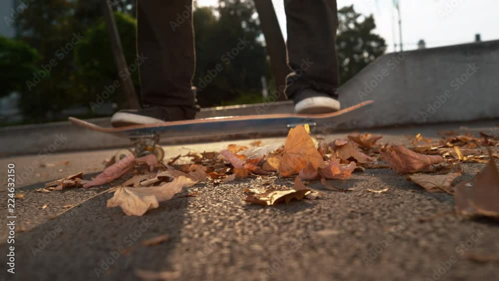 SLOW MOTION, CLOSE UP, LENS FLARE: Unrecognizable skater scatters the fallen autumn leaves around the asphalt pavement with his board after landing a trick. Cinematic shot of colorful leaves on road. SLOW MOTION, CLOSE UP, LENS FLARE: Unrecognizable skater scatters the fallen autumn leaves around the asphalt pavement with his board after landing a trick. Cinematic shot of colorful leaves on road.