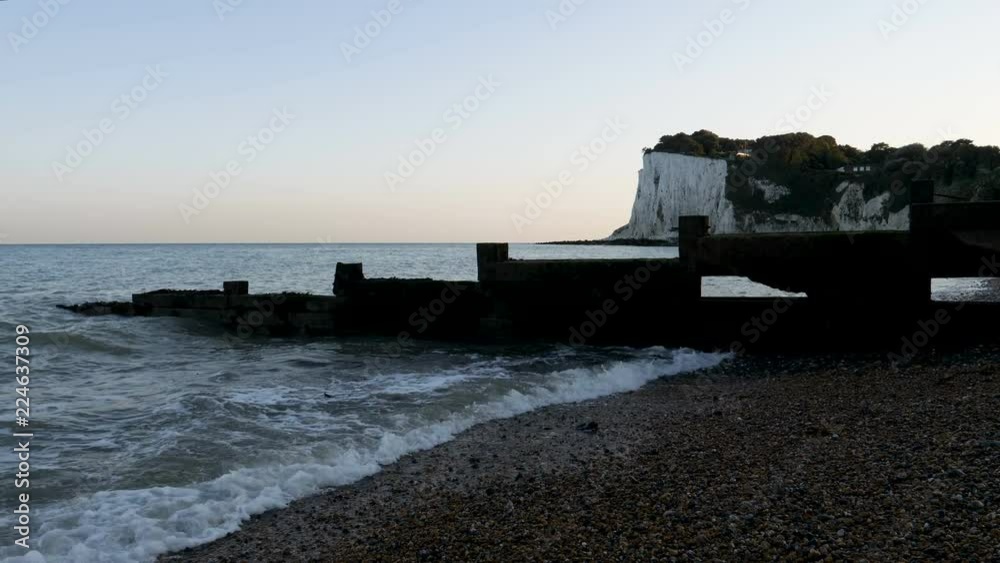 Waves wash onshore in the evening with seawalls in silhouette against the dark shadowy Dover cliffs in background Waves wash onshore in the evening with seawalls in silhouette against the dark shadowy Dover cliffs in background