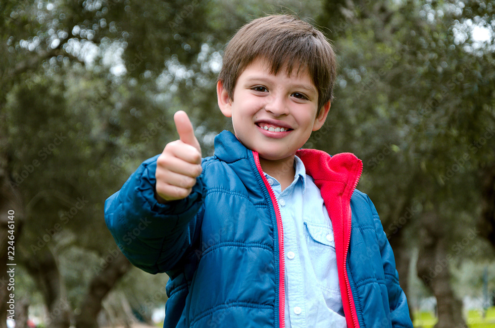 Portrait of a cute blond-haired boy, thumbs up and smile, with green background in the park Portrait of a cute blond-haired boy, thumbs up and smile, with green background in the park