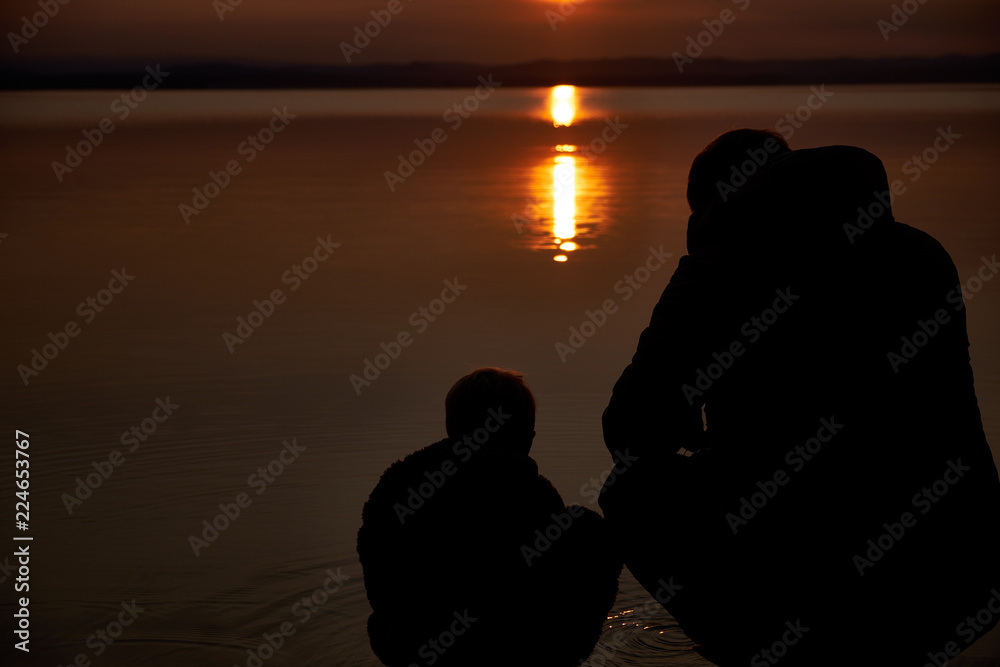 Silhouettes of father and his little son sitting together on the beach at sunset. Outdoors. Lifestyles concept.