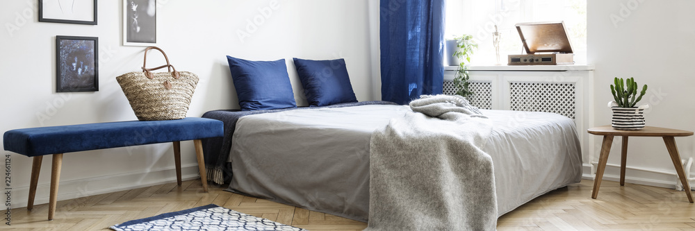 Real photo of a minimalistic bedroom interior with a bench next to a bed, wicker bag and pillows