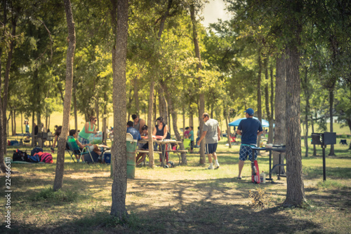 Fototapeta Naklejka Na Ścianę i Meble -  Blurred large group of friend, family members enjoy barbecue on lakeside area. Outdoor camping at natural park with picnic table in Grand Prairie, Texas, USA. Outside Party and BBQ Concept