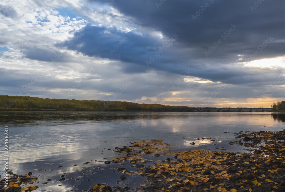 Panoramic picture of a calm river with blue waters, on which the yellow leaves lay, the sky is covered with clouds through which a ray of light tries to penetrate.