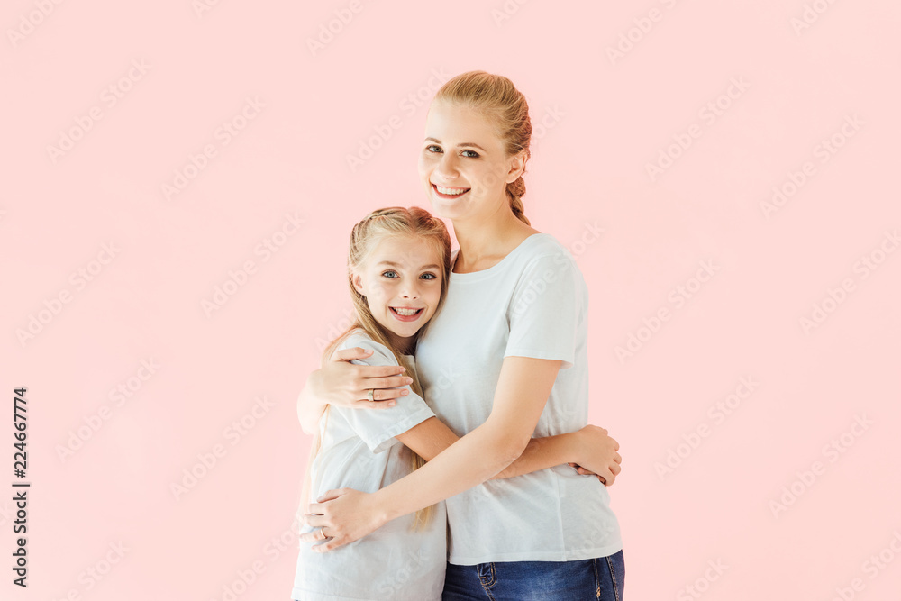 beautiful mother and daughter in white t-shirts embracing and looking at camera isolated on pink