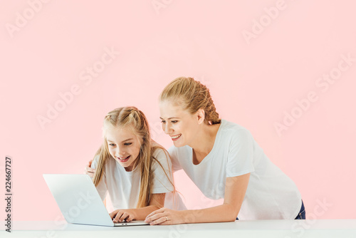 smiling mother and daughter in white t-shirts using laptop together isolated on pink