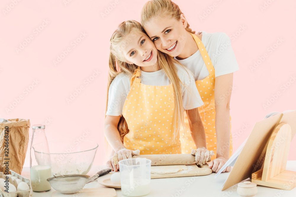 happy young mother and daughter kneading dough with rolling pin together and looking at camera isolated on pink