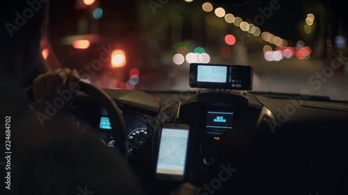 Taxi driver drives cab through night city streets or highway road, uses smartphone for navigation or uses shared economy phone app to find passengers or clients, taximeter counts fare