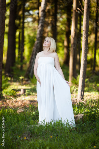 Beautiful pregnant woman in sheer long white maternity dress looking dreamy in the sky in the middle of forest