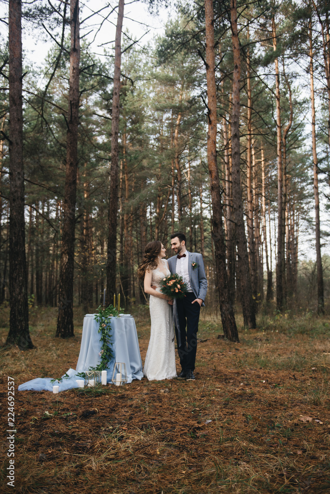 Fototapeta premium Happy bride and groom stand near the table, set for two in the woods. Autumn. The concept of a romantic date