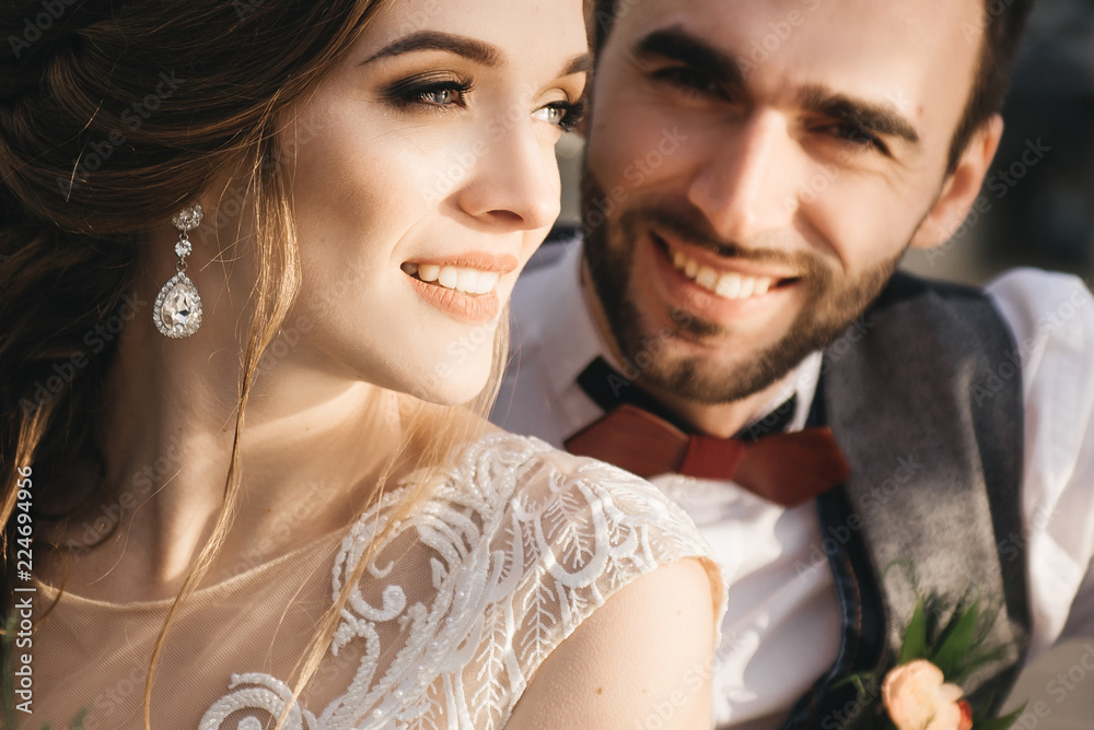 Happy young smiling bride and groom are sitting on the suspension bridge. Sunny wedding photos in an interesting place