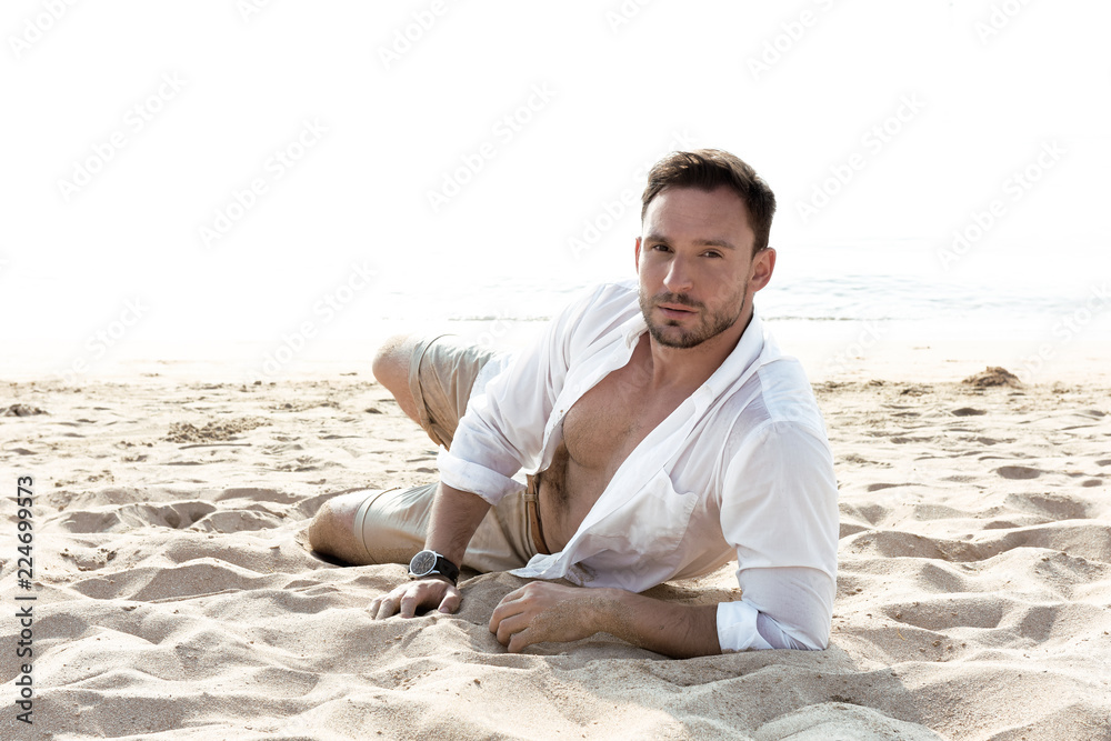 Handsome man in white shirt standing on a beach looking hot on summer day enjoying life. Brutal Male model poses with wet short in water naked chest out, smiling with smirk on his look and kind eyes.
