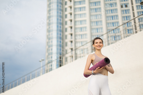 Young joyful woman in sporty top and white leggings holding purple yoga mat in hands while happily looking aside with skyscraper on background © Anton