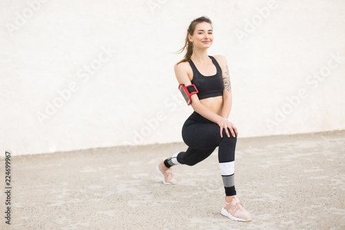 Beautiful smiling girl in dark gray sporty top and leggings with cellphone in armband happily looking in camera while doing sport exercises isolated
