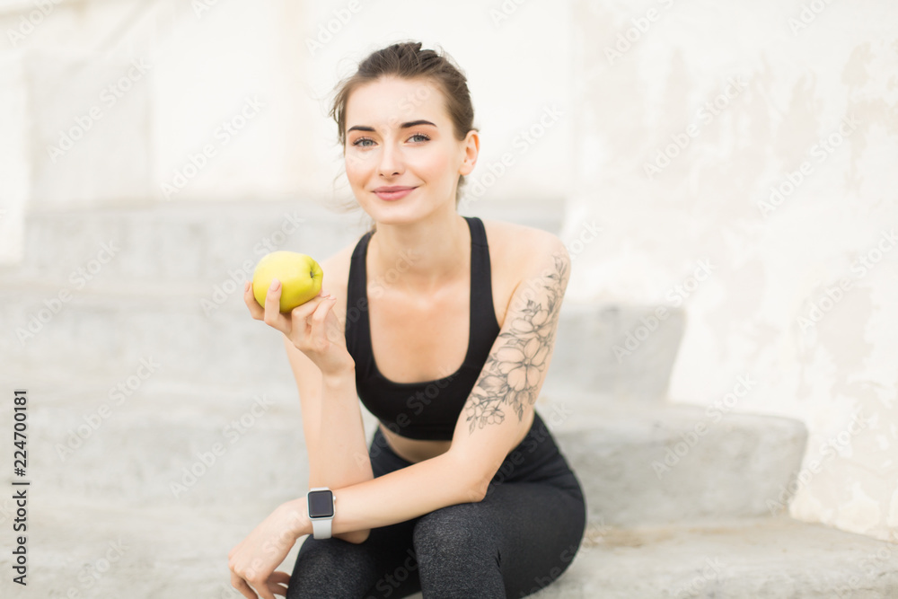 Young beautiful woman in sporty top and leggings holding apple in hand dreamily looking in camera while sitting on stairs outdoor Young beautiful woman in sporty top and leggings holding apple in hand dreamily looking in camera while sitting on stairs outdoor