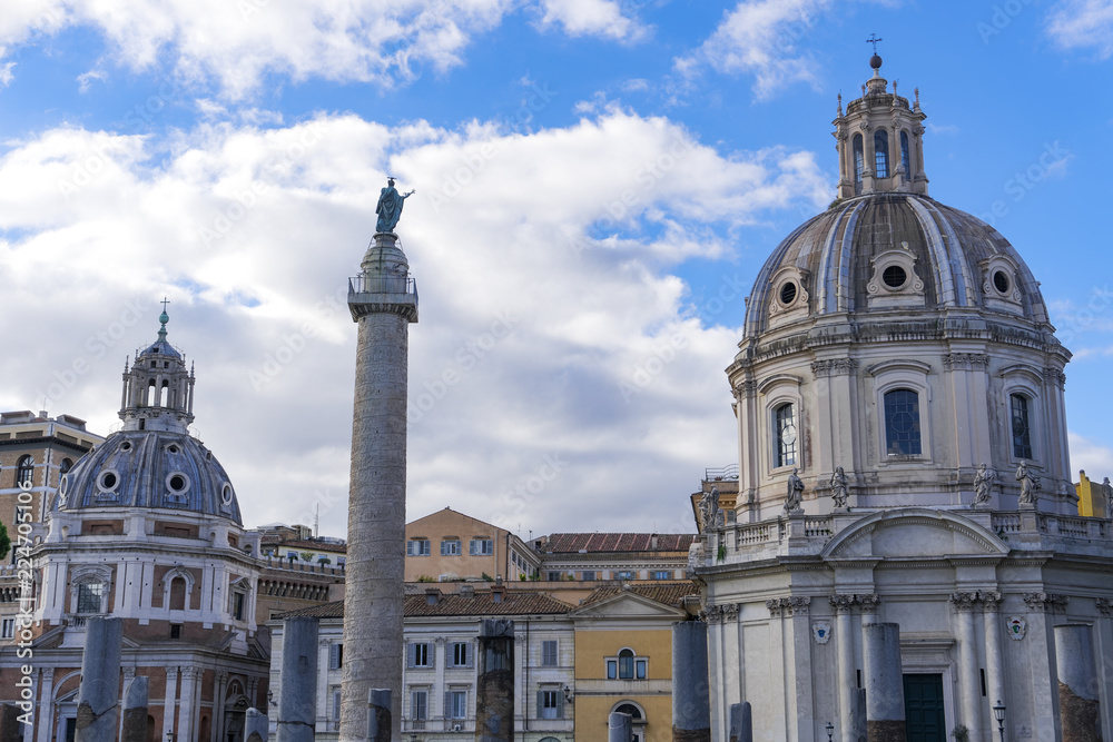 Rome, Italy Trajan triumphal column with top viewing platform on the Roman Forum. Freestanding marble Colonna Traiana on pedestal with background view of Roman Catholic churches rooftops.