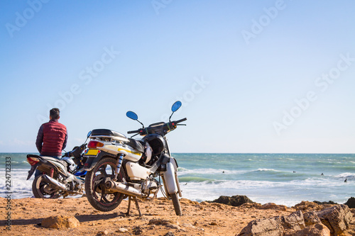 A guy in a burgundy waistcoat on a motorcycle against the background of the ocean. The coast of the Atlantic Ocean. The beach is near Agadir. Africa Morocco