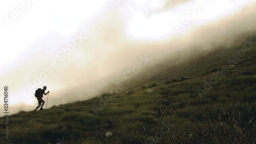 Young trekker man walking up the mountain in the fog with backpack, boots and trekking poles. Clouds and myst moving on the background. Abruzzo Italy.