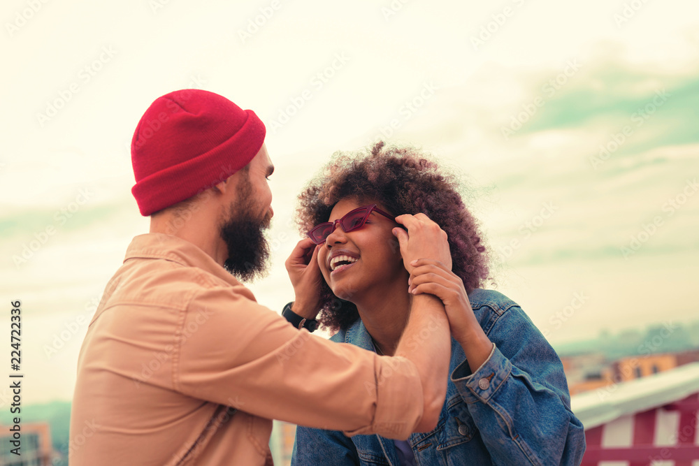 New glasses. Careful young man looking at his smiling beautiful girlfriend while helping her with wearing red glasses New glasses. Careful young man looking at his smiling beautiful girlfriend while helping her with wearing red glasses