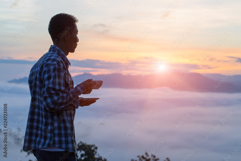 Fototapeta premium A tourist holding a cup of coffee on the mountain,The valley is covered with fog and has a beautiful sunrise. Beauty of nature, tourism and traveling concept.