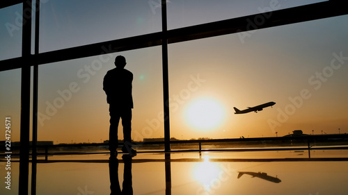 фотографія Silhouette of a tourist guy watching the take-off of the plane standing at the airport window at sunset in the evening
