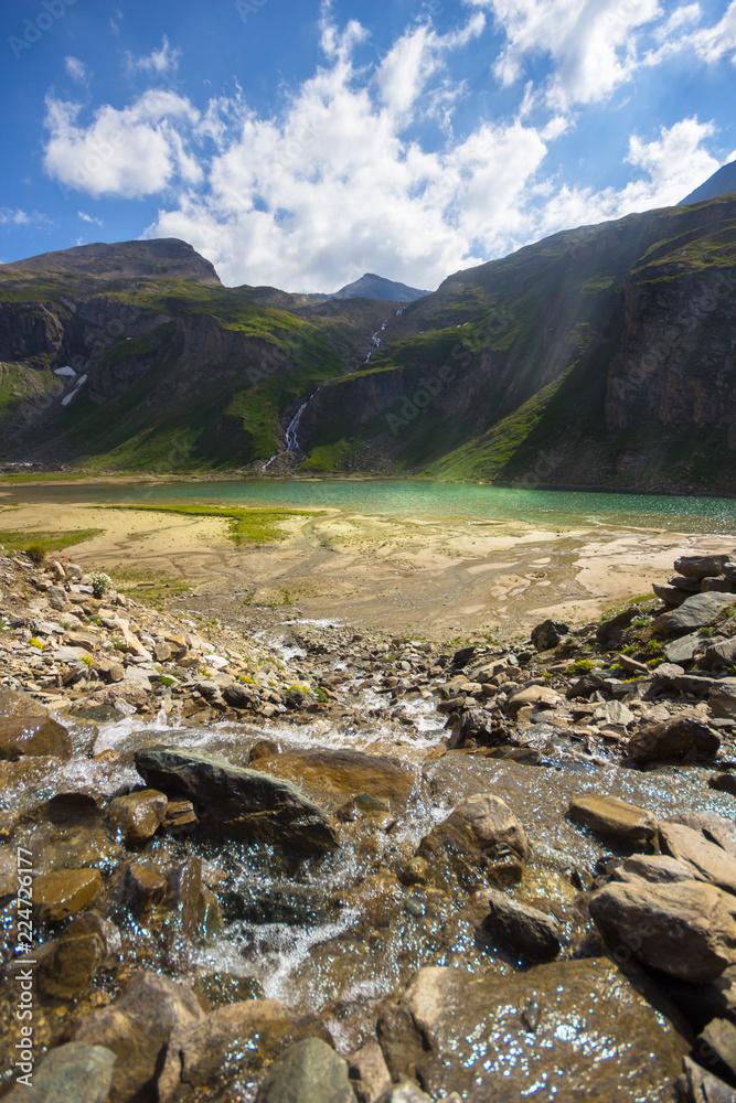 Standing in the center of a wild mountain creek, flowing in rivulets into the Nassfeldspeicher Lake, a so called power place, with sun rays enlightening the water surface, in the High Tauern, Austria