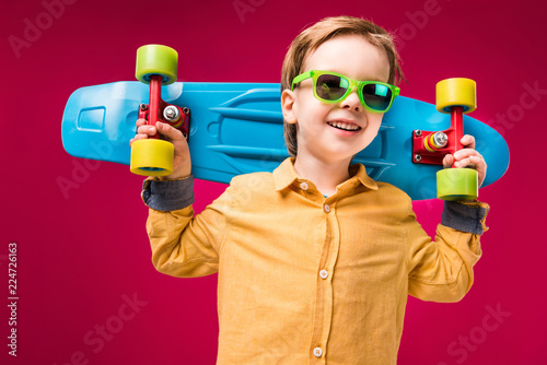 Stylish smiling boy in sunglasses posing with penny board isolated on red