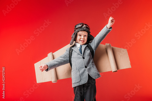little emotional pilot in suit and cardboard plane wings with outstretched arms to fly isolated on red