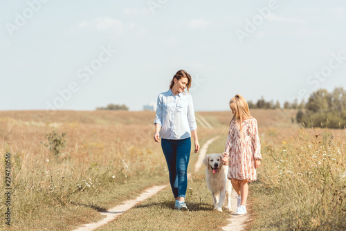 happy mother and daughter walking with golden retriever in field
