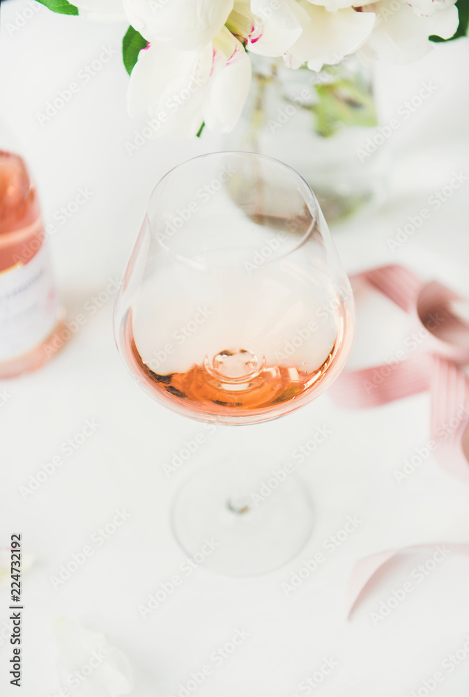 Rose wine in glass, pink decorative ribbon, peony flowers over white background, selective focus, close-up. Summer celebration, wedding greeting card, invitation concept