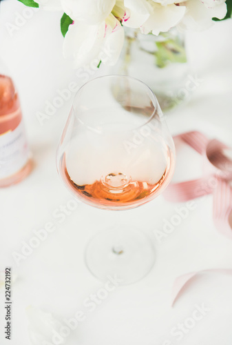 Rose wine in glass, pink decorative ribbon, peony flowers over white background, selective focus, close-up. Summer celebration, wedding greeting card, invitation concept