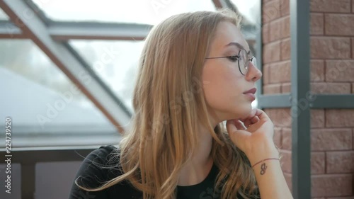 Young beautiful girl in glasses sitting in a cafe by the window looking at the sides