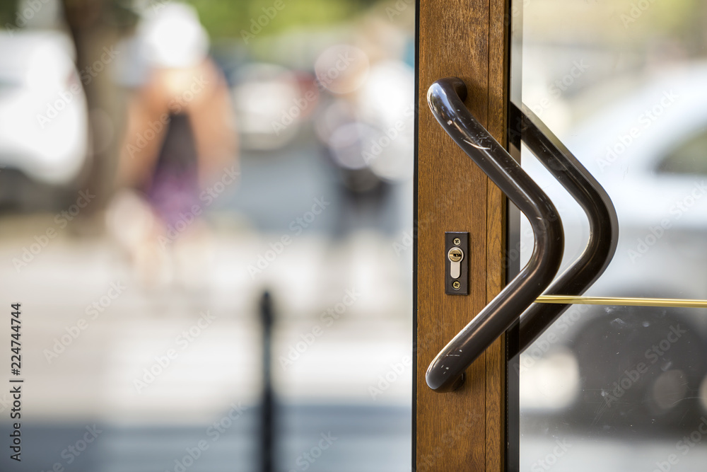 Fototapeta premium Close-up of plastic or wooden modern brown door with transparent glass surface and big metal handle on blurred bright copy space bokeh background. Entrance, exit, office or hallway.