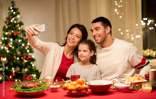 holidays, family and celebration concept - happy mother, father and little daughter having christmas dinner and taking selfie picture by smartphone at home