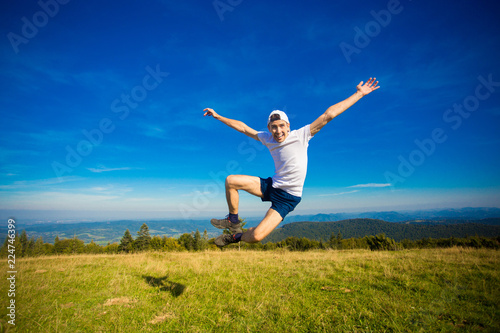 Summer hiking in mountains. Young tourist man in cap with hands up on top of mountains admires nature. Travel concept