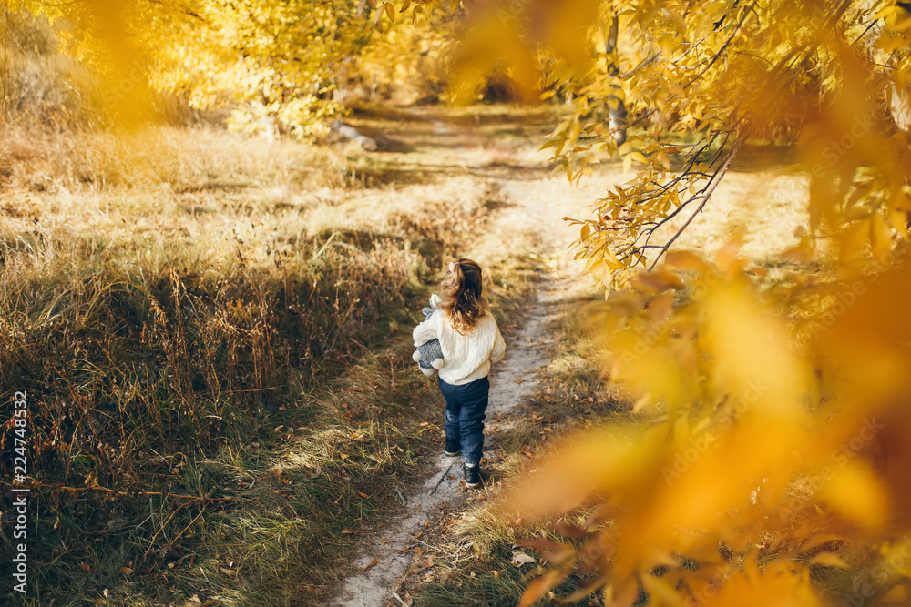 Back view of trendy girl running from his mother while playing outdoor. Toddler enjoying sunny weather and autumn sunshine. Lifestyle concept Back view of trendy girl running from his mother while playing outdoor. Toddler enjoying sunny weather and autumn sunshine. Lifestyle concept