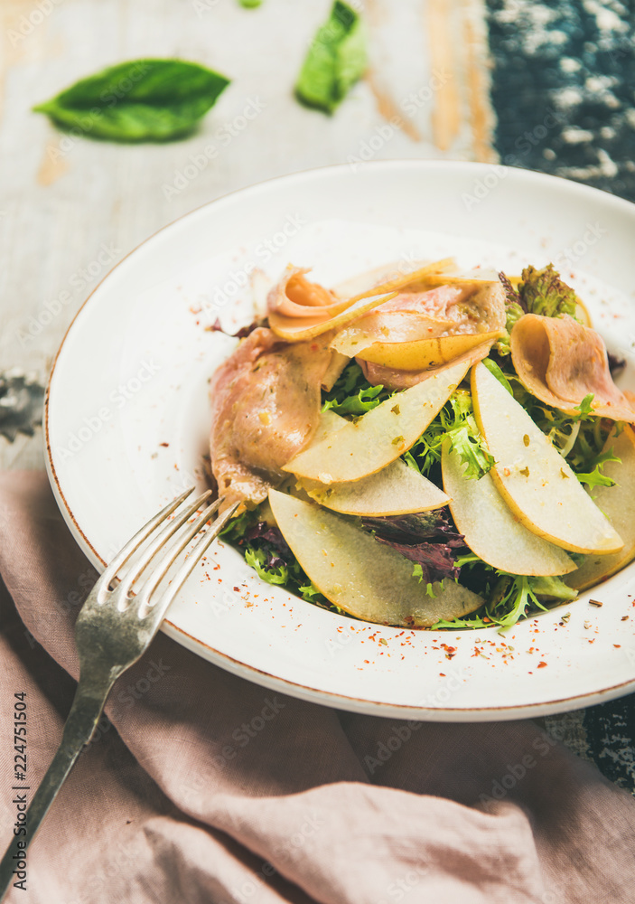 Fresh summer salad with smoked turkey ham and slices of pear in white plate over scorched rustic wooden table background, selective focus, vertical composition
