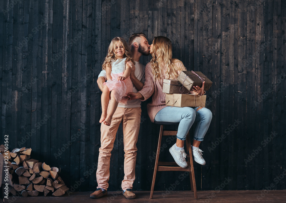 Family concept. Handsome father holds daughter on hands and kiss his wife with gifts in empty room against a wooden wall.