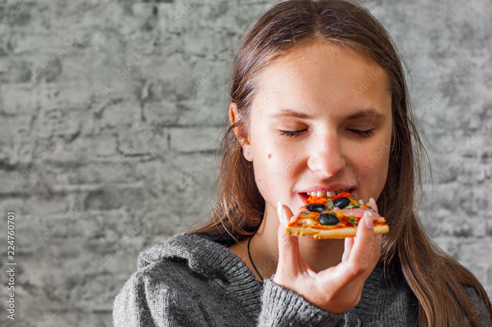 portrait of young teenager brunette girl with long hair eating slice of pizza on gray wall background