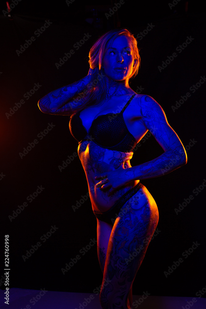 Muscled caucasian woman with tattoos and oiled skin poses under colored lights