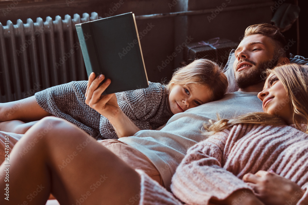 Mom, dad and daughter reading storybook together while lying on bed. - obrazy, fototapety, plakaty 