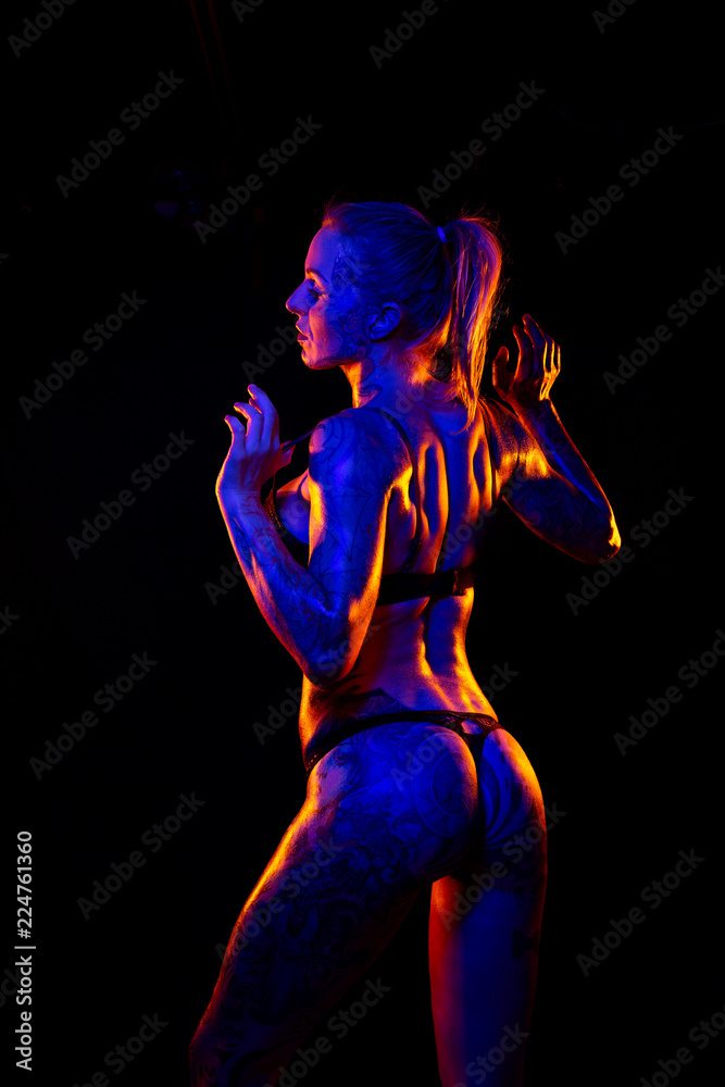 Fototapeta premium Muscled caucasian woman with tattoos and oiled skin poses under colored lights
