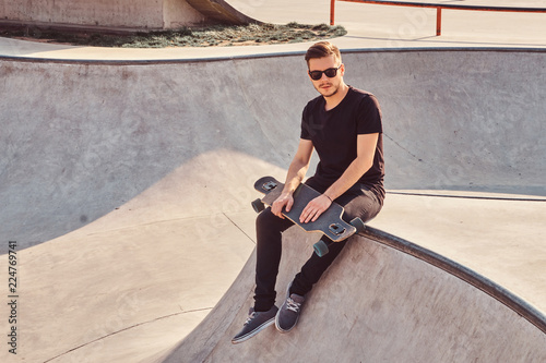 Stylish hipster in sunglasses dressed in black jeans and shirt holds skateboard while sitting on skatepark. © Fxquadro