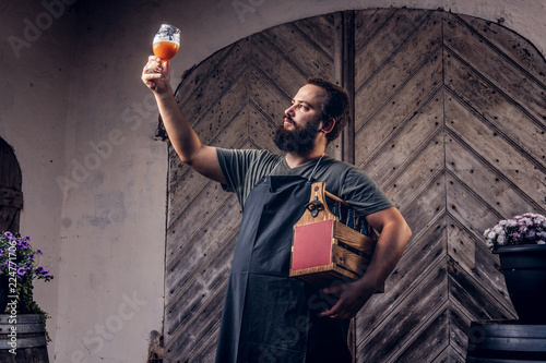 Expert brewer in apron holds glass of beer and checking quality of brewed drink at brewery factory.