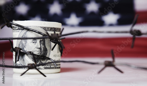 US Dollar money wrapped in barbed wire on United States national flag background as symbol of economic warfare, sanctions and embargo busting