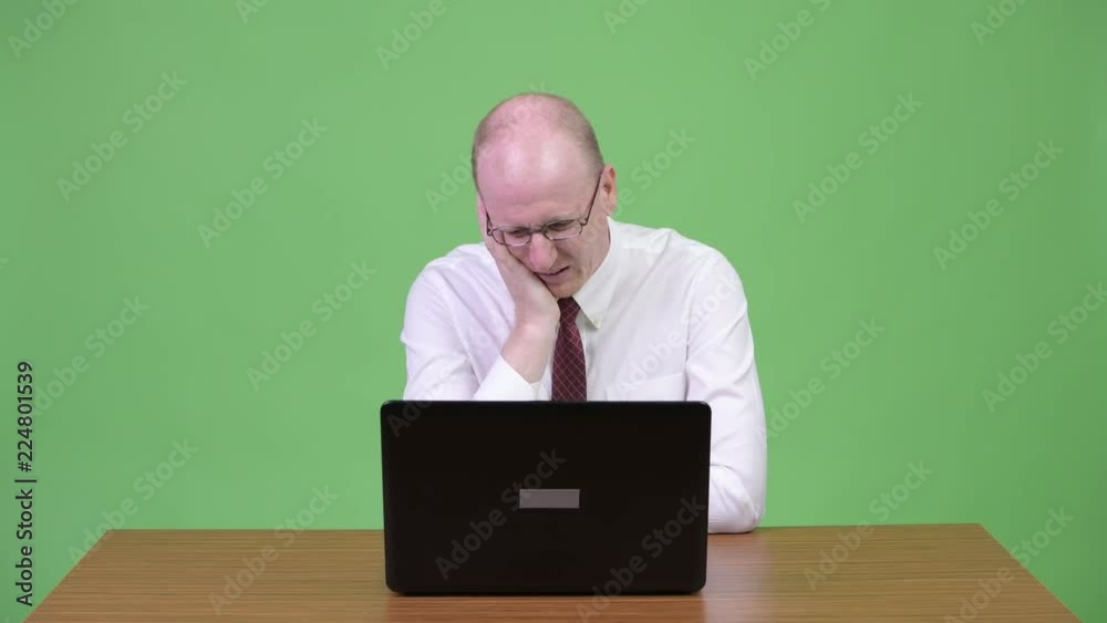 Stressed mature bald businessman using laptop and getting bad news against wooden table Stressed mature bald businessman using laptop and getting bad news against wooden table