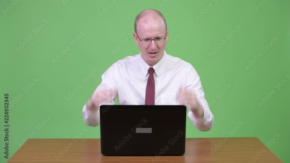Angry mature bald businessman talking to the laptop against wooden table Angry mature bald businessman talking to the laptop against wooden table