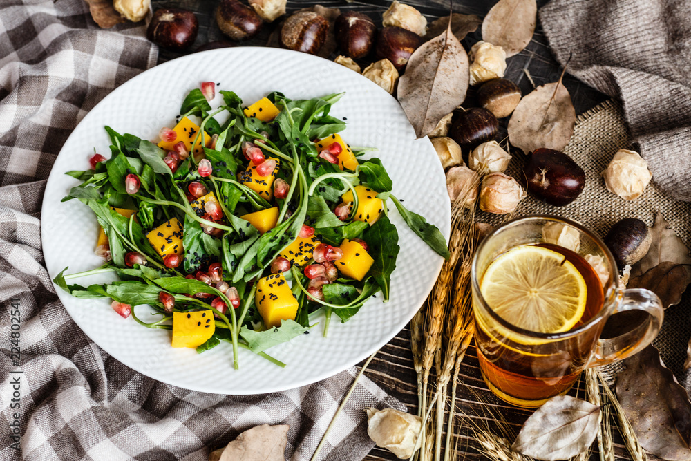 Food flat lay. Top view. Salad with mango, arugula, pomegranate seeds lying on white plate. Next lying pumpkin, cup of black tea with lemon, dry flowers and leaves, physalis, chestnuts, sweater Food flat lay. Top view. Salad with mango, arugula, pomegranate seeds lying on white plate. Next lying pumpkin, cup of black tea with lemon, dry flowers and leaves, physalis, chestnuts, sweater