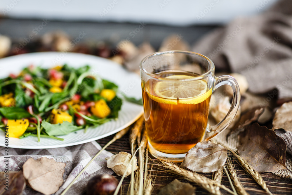 Salad with mango, arugula, pomegranate seeds lying on white plate. Next lying pumpkin, cup of black tea with lemon, dry flowers and leaves, physalis, chestnuts, sweater Salad with mango, arugula, pomegranate seeds lying on white plate. Next lying pumpkin, cup of black tea with lemon, dry flowers and leaves, physalis, chestnuts, sweater